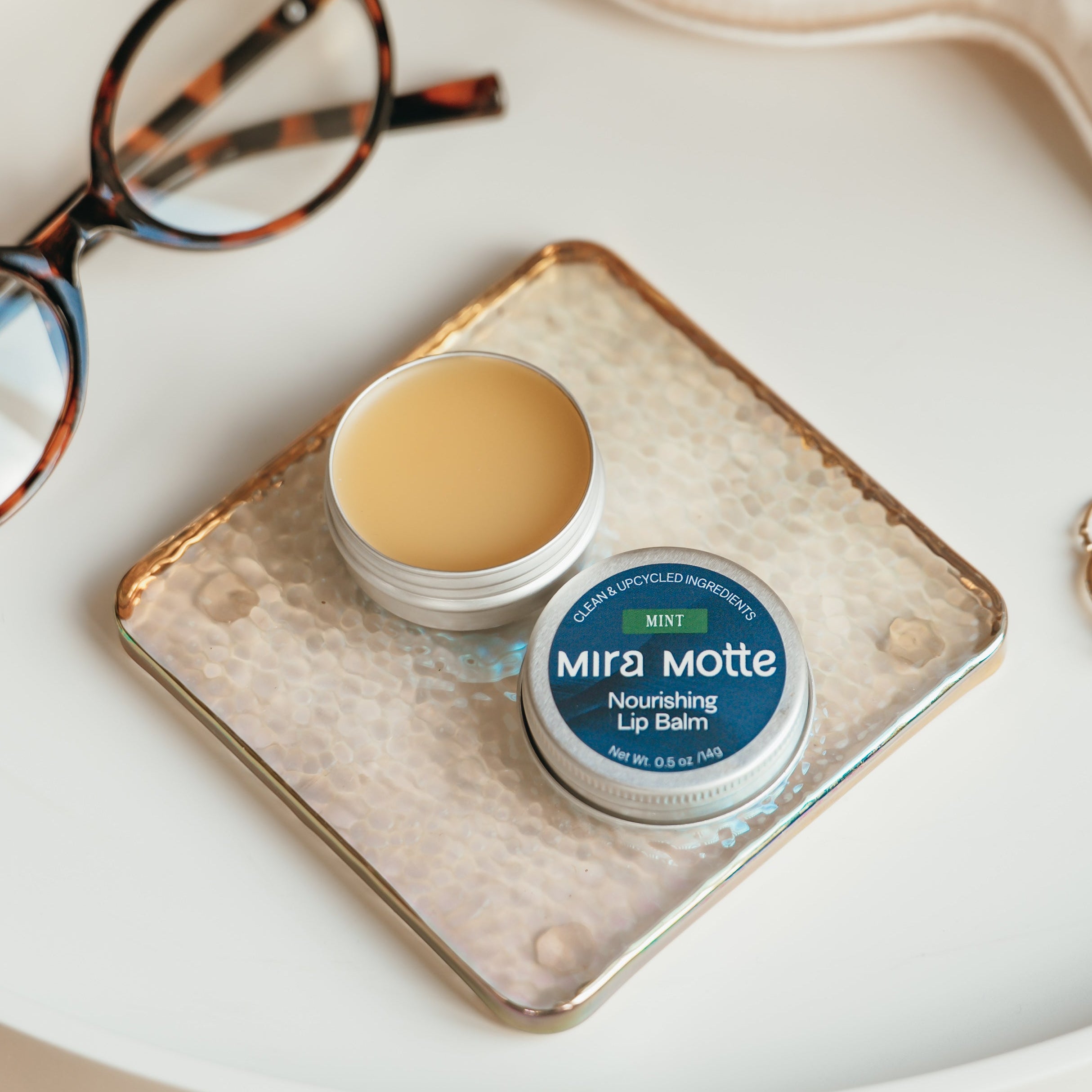 Mira Motta lip balm on a coaster with glasses, ring, and face mask on a table