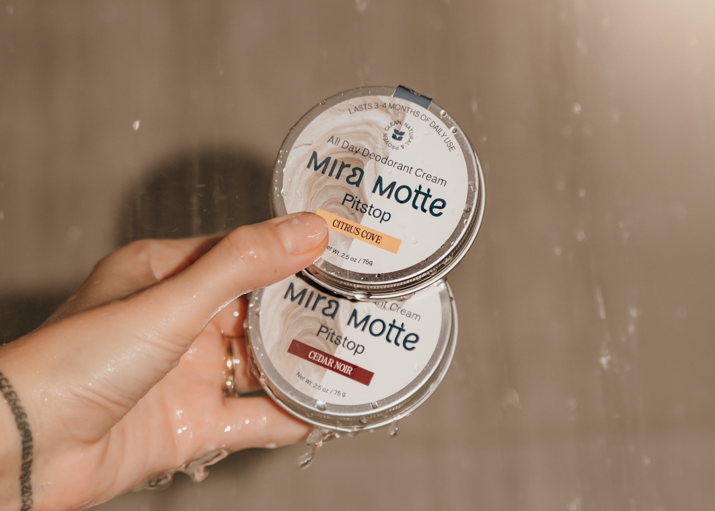 Hand holding two containers of Mira Motte Pit Stop cream against a blurred background