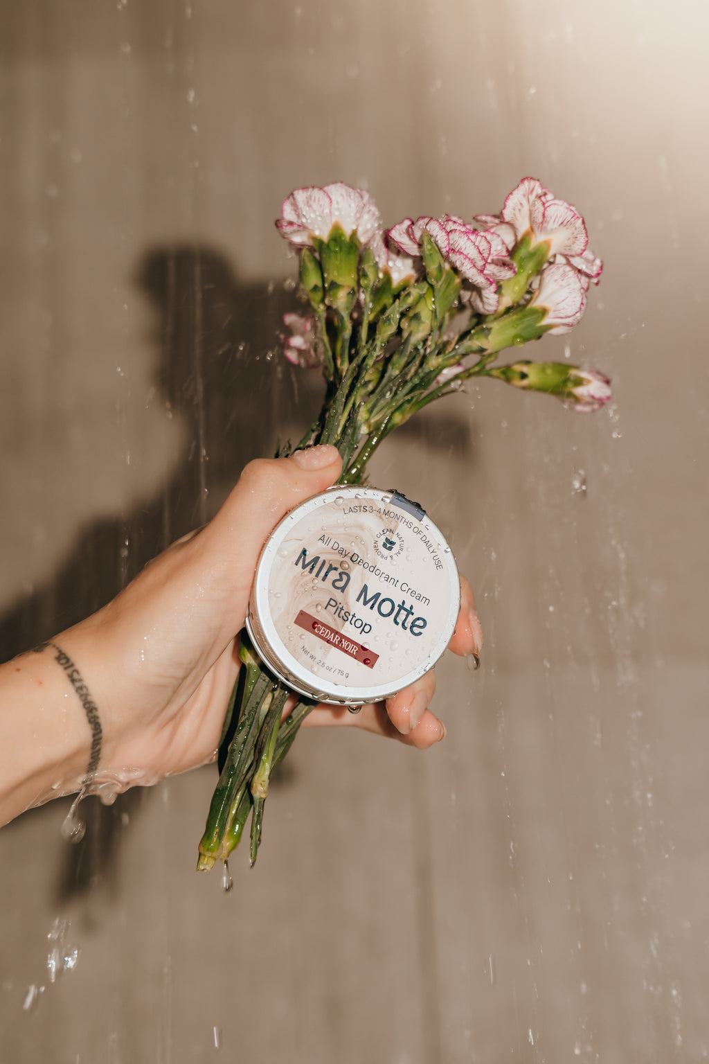 Hand holding a small container labeled 'Mira Motte' with a bouquet of flowers against a neutral background.