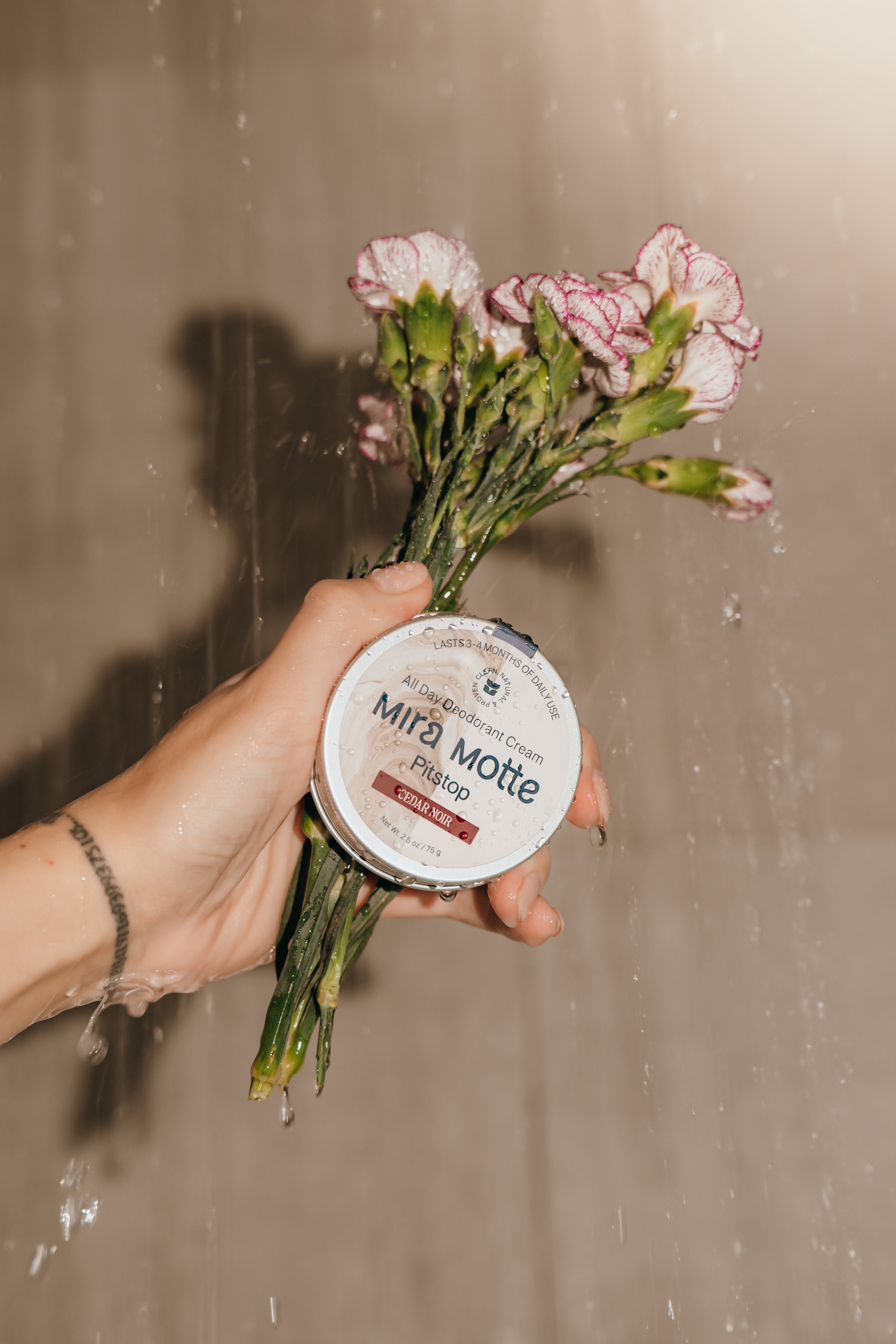 Hand holding a small container labeled 'Mira Motte' with a bouquet of flowers against a neutral background.