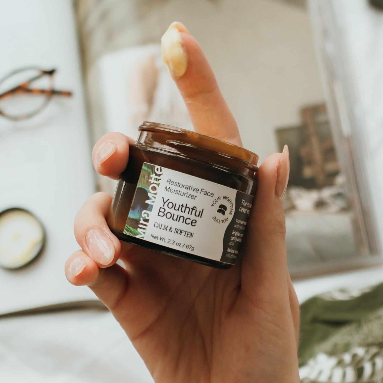 Hand holding a jar of 'Youthful Bounce' face cream with a blurred indoor background.