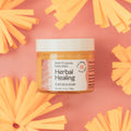 Jar of Herbal Healing balm on a pink background with yellow flowers