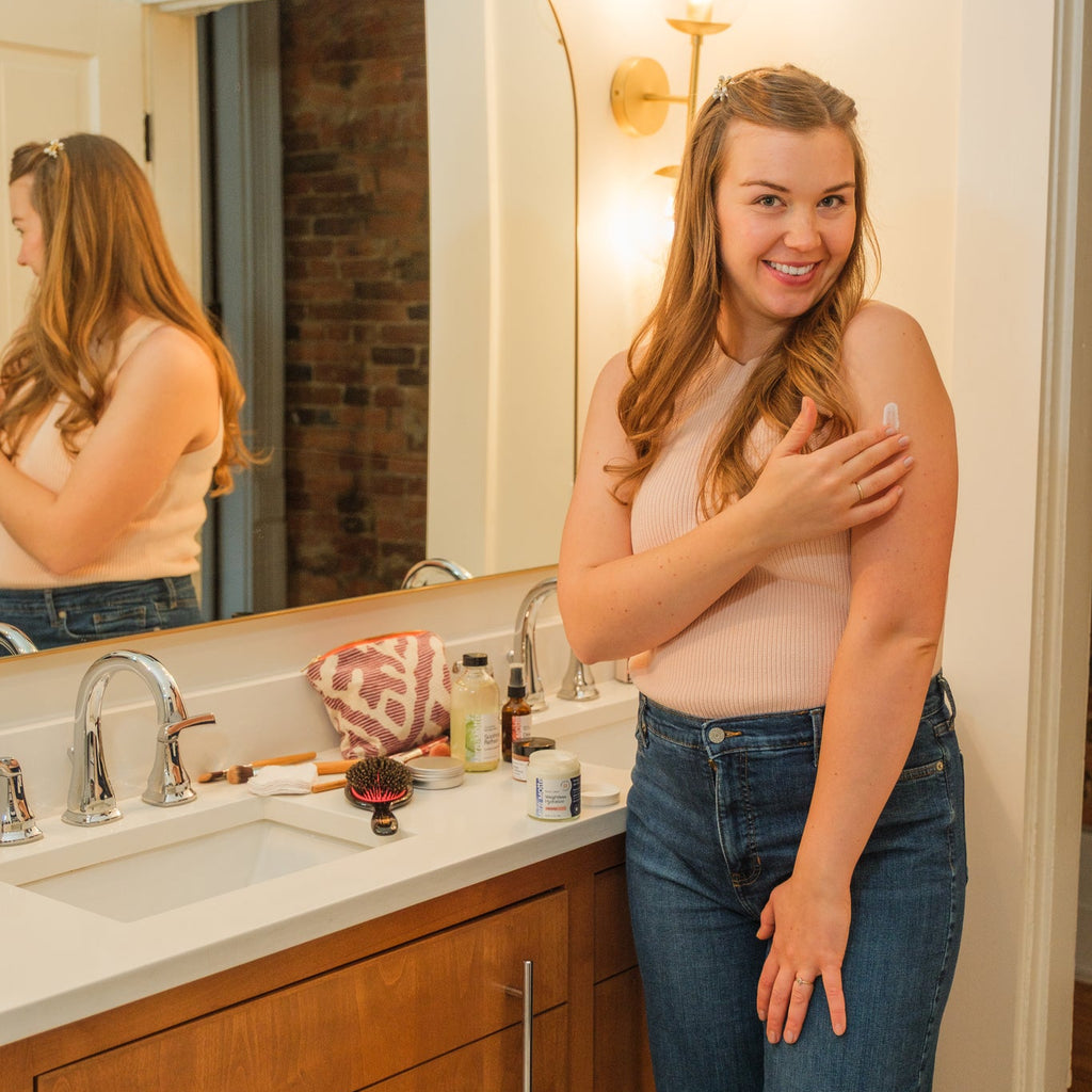Woman standing in a bathroom applying lotion to her arm