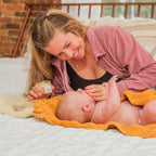 Woman lying on a bed putting baby balm on a baby on an orange blanket, both smiling.