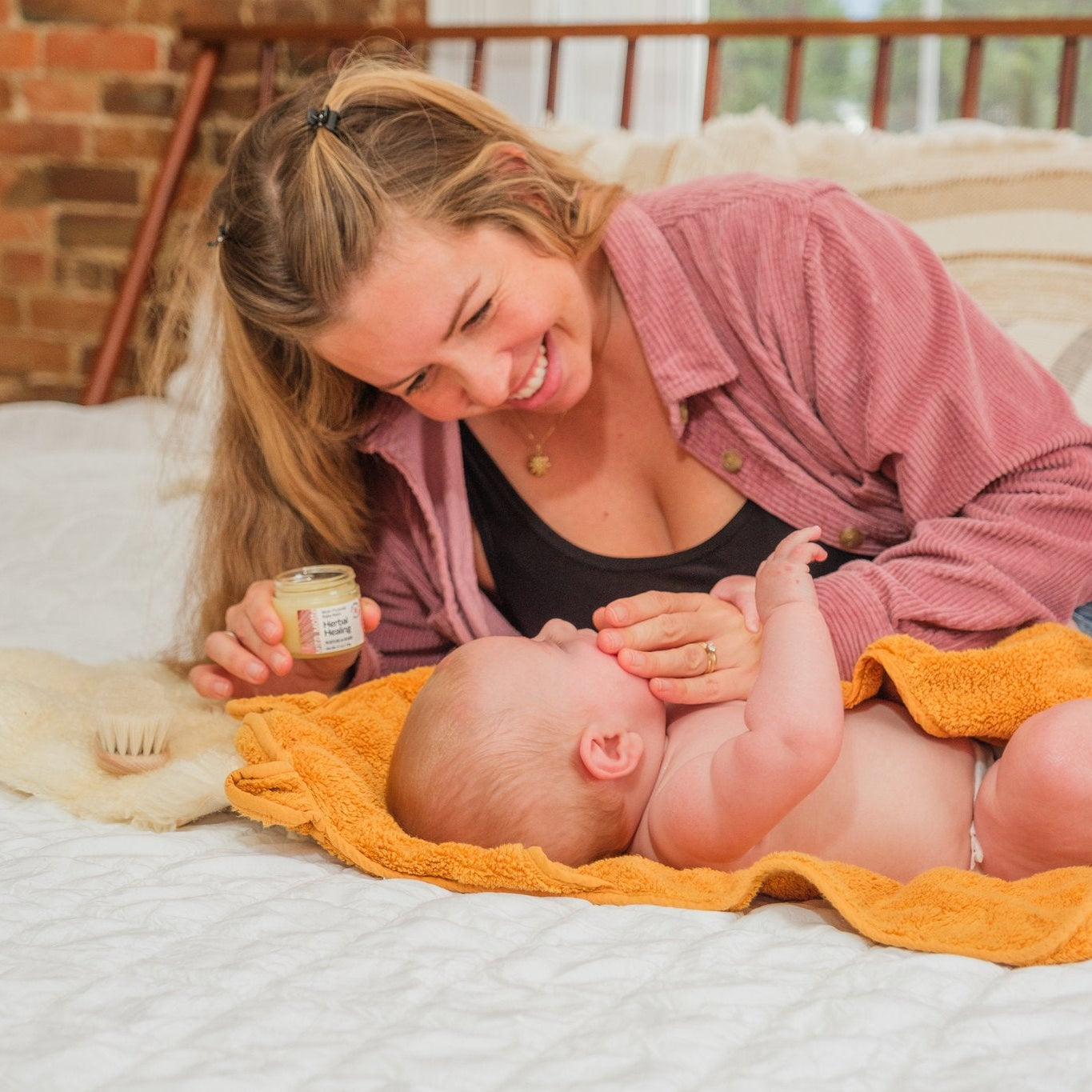Woman lying on a bed putting baby balm on a baby on an orange blanket, both smiling.