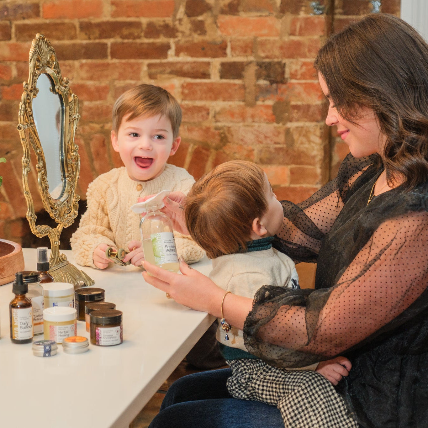Woman with two children in a room with a brick wall and a mirror. Woman is applying Mira Motte micellar water to a reusable cotton pad. 
