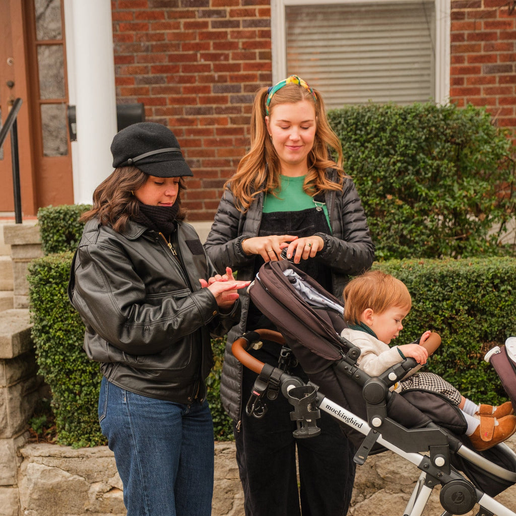 Two women with a child in a stroller on a sidewalk applying hand salve. 